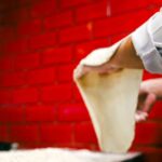 Chef skillfully stretches pizza dough in a vibrant kitchen with red brick walls. - Photo by Yusuf Çelik on Pexels
