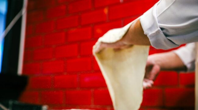 Chef skillfully stretches pizza dough in a vibrant kitchen with red brick walls. - Photo by Yusuf Çelik on Pexels